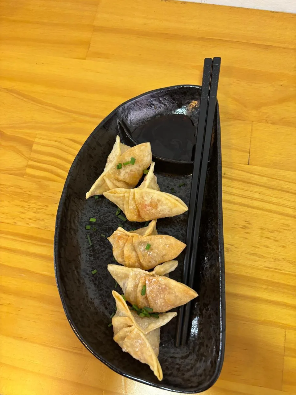 Gyozas doradas con cebollín y salsa en un plato negro sobre una mesa de madera