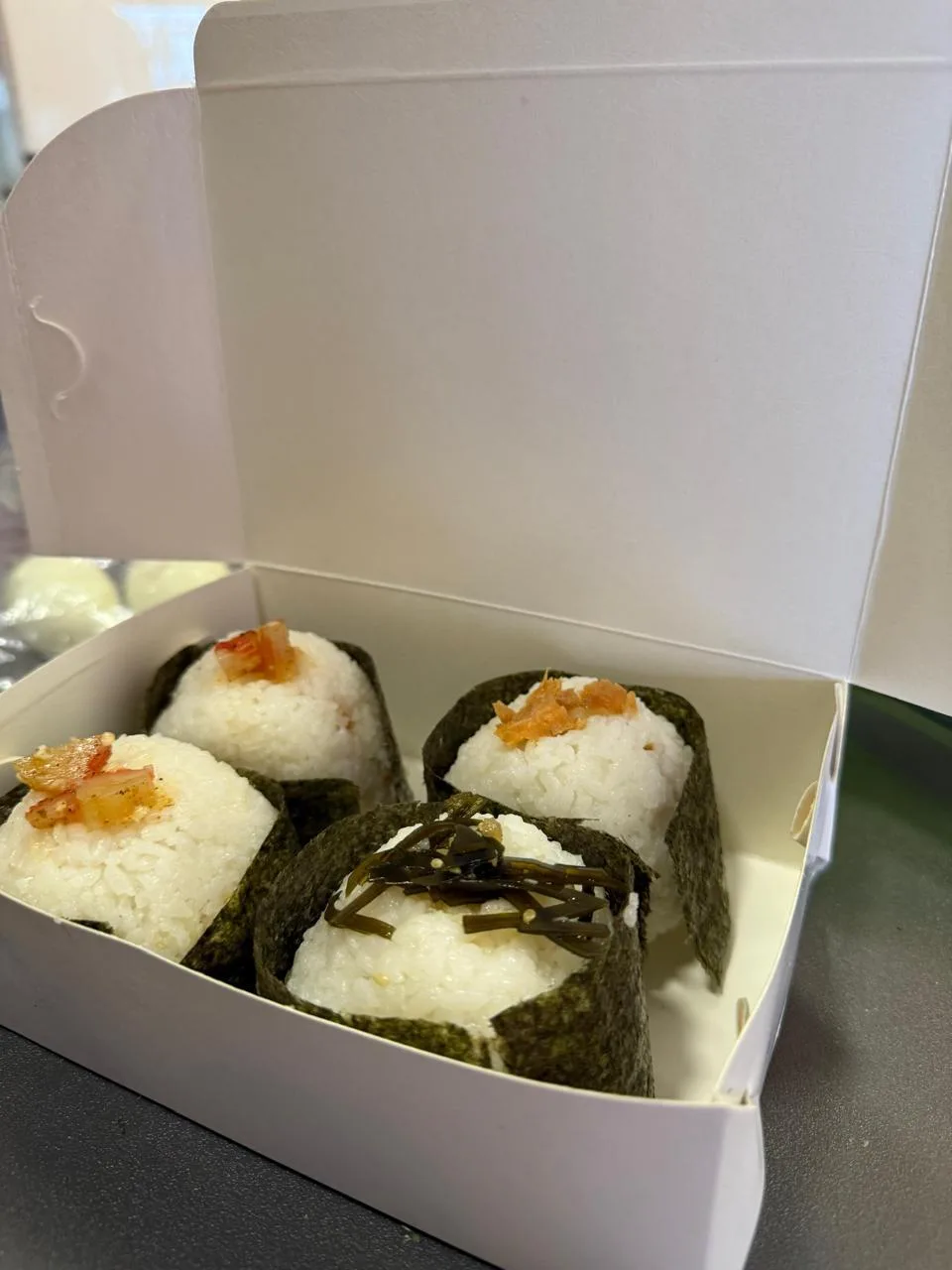 Cuatro onigiri envueltos en alga nori, cada uno con un ingrediente distinto encima, dentro de una caja
